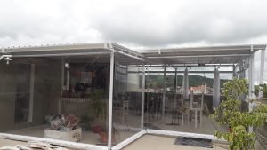 Cobertura de Telha Sanduiche, pintada em branca, com estrutura em aço galvanizado e pin com fechamentos com cortinas rolô, de visor e bordas brancas.